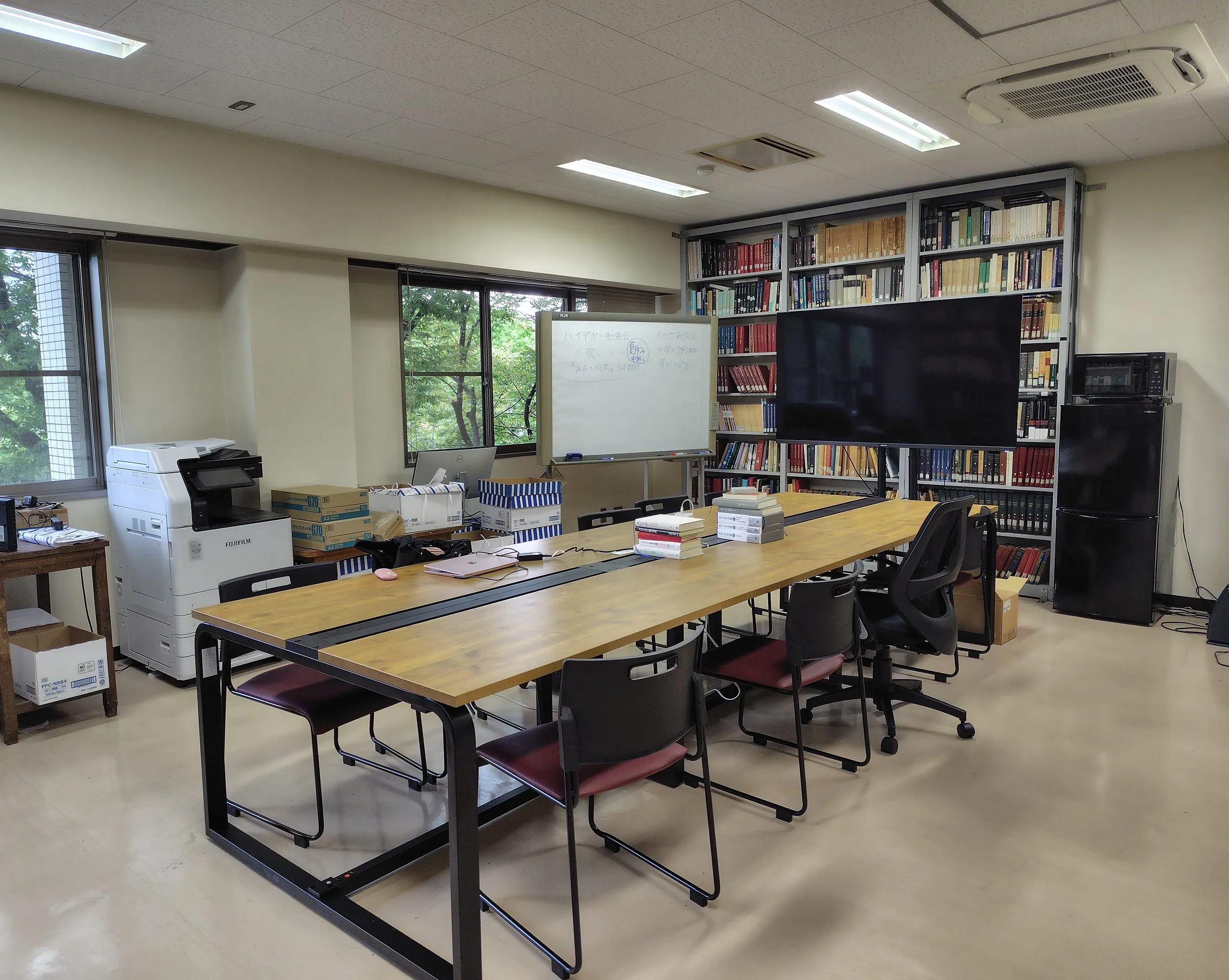 哲学リテラチャーラボ（311号室）。ホワイトボードと大型モニタ、本棚に囲まれた木の長机